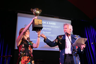 Photo of FVAP's Executive Director, Erin Smith, holding the Battle of the Band Trophy up on stage next to the emcee. 