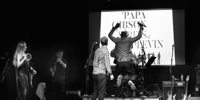 Papa Gibson singer jumping on stage with mic in hand and band logo in background.